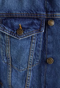 Blå denimskjorte med spiss lomme, brune knapper og gule sømmer. Teksturert stoff med synlige vevdetaljer.