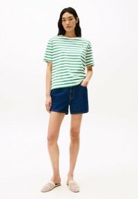 Camiseta rayas verde y blanca, de corte relajado, combinada con pantalones cortos de mezclilla azul oscuro. El modelo lleva puestos unos zapatos de lona beige sin cordones. Diseño simple, look casual.