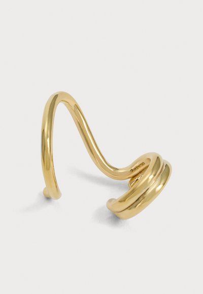 Manchette d’oreille sculpturale de ton doré, avec une bande incurvée et trois boucles arrondies, présentée sur un fond clair uni.