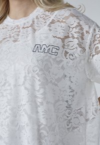 Hvid bluse i lace med blomsterdesign, rund halsudskæring og broderet "NYC" logo i marineblå. Den transparente struktur giver en let og luftig fornemmelse.