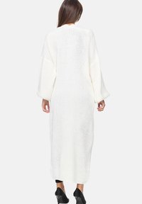 Lång, vit, stickad cardigan med lös passform, vida ärmar och en texturerad mönstring, som når till mitten av vaden. Kombineras med svarta byxor och skor.