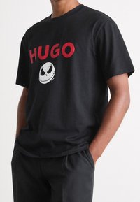 Musta puuvillainen t-paita, jossa on punainen "HUGO" -teksti ja valkoinen Jack Skellington -grafiikka. Pyöreä kaula-aukko, lyhyet hihat, rento malli.
