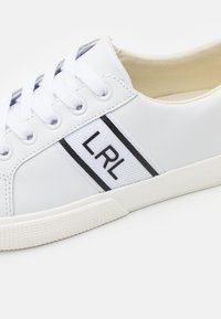 Basket blanche basse avec lacets blancs et une bande en tissu sur le côté affichant des lettres noires "LRL" et deux lignes noires verticales.