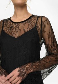 Černý krajkový top s průsvitnými dlouhými rukávy, květinovými vzory a výstřihem do V přes jednobarevnou černou vrstvu. Texturované krajkové detaily.