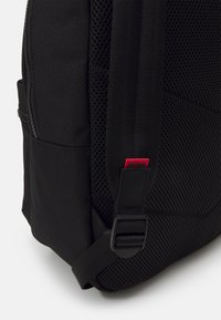 Sac à dos noir avec tissu durable, panneaux en mesh, bretelles réglables et un logo rouge en accent. Compartiment zippé sur le côté.
