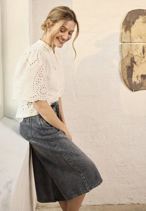 Noor naine blondide juustega valges silmahõlmadega pluusis ja hallis teksaseelikus istub naeratades valge seina ääres, millel on abstraktne seinakunst.