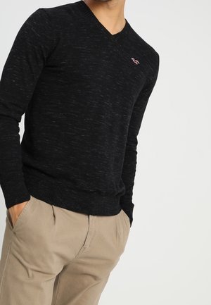 Homme portant un pull noir à col en V avec un petit logo sur la poitrine et un pantalon beige, les mains dans les poches, sur fond blanc uni.