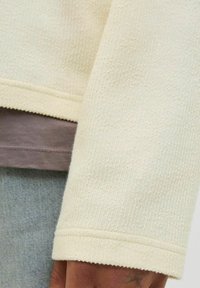 Manche de pull texturé crème avec un tissage épais, dotée d'un poignet simple et superposée à un haut gris clair, associée à un denim.