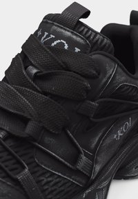 Κοντινό πλάνο μαύρου αθλητικού παπουτσιού με χοντρές υφασμάτινες κορδόνια και το λογότυπο «KOI» στο πλάι και στη γλώσσα.