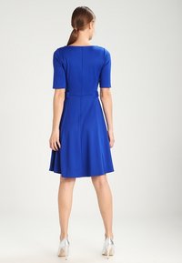 Robe A-line bleue avec des manches courtes, une finition en tissu lisse et une taille marquée. Présente une fermeture éclair au dos et de subtils plis à l'ourlet.