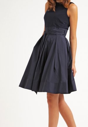 Sort ærmeløs kjole med tætsiddende krop og plisseret, knælængde skirt med flæse og bindebånd ved taljen.