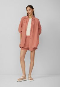 Leichtes, strukturiertes Hemd in rostfarben mit Knopfverschluss, kombiniert mit passenden Shorts und weißen Sandalen mit Riemchen. Einfaches und stimmiges Design.