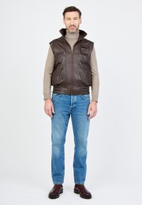 Braune Lederweste mit hohem Kragen, Reißverschluss und mehreren Taschen, kombiniert mit hellblauen Jeans und dunkelbraunen Stiefeln.