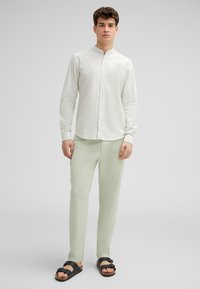 Leichte, strukturierte Creme-Blusen mit Stehkragen, kombiniert mit hellgrünen Hosen und schwarzen Sandalen mit einer Schnallen-Detail.