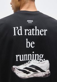 Άτομο που φοράει μαύρο μπλουζάκι Adidas με το κείμενο «Θα προτιμούσα να τρέχω.» και εικόνα ενός λευκού παπουτσιού τρεξίματος Adidas στην πλάτη.
