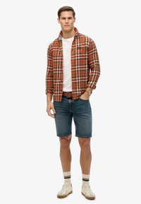 Camisa de franela a cuadros en naranja y verde, camiseta blanca, pantalones cortos de mezclilla y zapatillas de color crema. Incluye calcetines a rayas y un reloj.