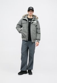 Graue Steppjacke mit fellgefütterter Kapuze, zwei Vordertaschen, schwarzes T-Shirt, graue Hose und schwarze Sneaker. Modell steht vor einem weißen Hintergrund.