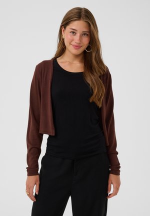 Junge Frau mit langen braunen Haaren, die goldene Creolen, einen braunen kurzen Cardigan, ein schwarzes Top und schwarze Hosen trägt, lächelt sanft.