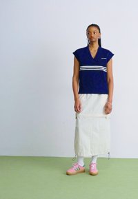 Σκούρο μπλε cropped τοπ με λευκές ρίγες, σε συνδυασμό με φούστα μακριά σε ανοιχτό μπεζ χρώμα. Το μοντέλο φοράει ροζ αθλητικά με πράσινες λεπτομέρειες και άσπρες κάλτσες.