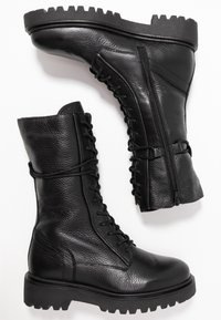 Pár černých kožených bojových bot s šněrováním v přední části, bočními zipy, texturovanými silnými podrážkami a kulatými špičkami na bílém pozadí.