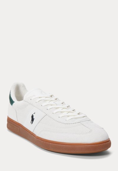 Witte suède sneaker met bruine rubberen zool, groene hieltab, witte veters en zwart polo-speler logo aan de zijkant.