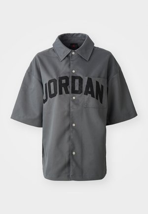 Pelēka īsām piedurknēm krekls ar pogu aizdari, kurā uz priekšpuses lieliem melniem burtiem ir rakstīts "JORDAN" un ir krūšu kabata. Gluda tekstūra.