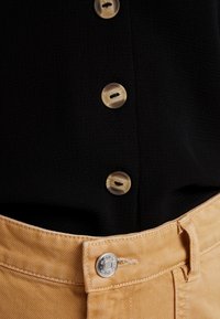 Camicia nera con texture e quattro bottoni marroni chiari a marmo, infilata in pantaloni beige di denim con un bottone metallico e passante per cintura.