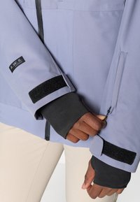 Violetti takki mustilla yksityiskohdilla, jossa on tarranauhateippaukset ja vetoketjullinen tasku. Teksturoitu kangas, joka on muotoiltu.