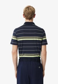 Polo shirt blu con righe bianche e verdi, maniche corte e colletto semplice; indossato con pantaloni scuri con una tasca posteriore e logo.