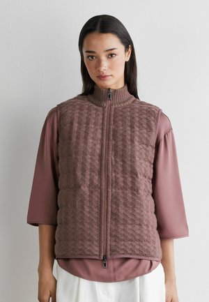 Vrouw draagt een mauve gewatteerd vest met rits over een blouse met driekwart mouwen en witte broek, staand tegen een effen lichte muur.