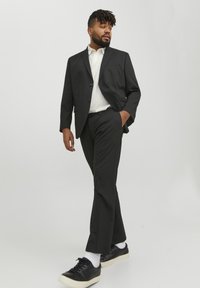 Traje negro con chaqueta de botonadura simple, pantalones ajustados y camisa blanca. Zapatillas negras con suelas blancas y calcetines blancos texturizados.