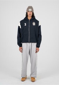 Námornícka mikina na zips s bielymi akcentmi na pleciach, logo na hrudi, skombinovaná so svetlošedými teplákmi so bočnými prúžkami a pieskovými topánkami.