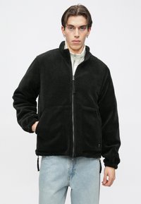 Casaco de lã polar preto com fecho de correr frontal, gola alta, dois bolsos frontais e punhos elásticos. Usado sobre uma t-shirt branca com jeans azul claro.