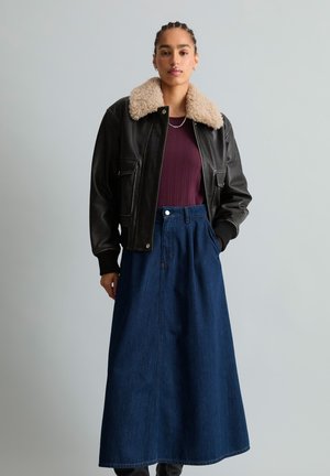 Junge Frau trägt eine schwarze Lederjacke mit einem beigen Pelzkragen, ein bordeauxfarbenes Oberteil und einen langen dunkelblauen Jeansrock, steht vor einem schlichten Hintergrund.