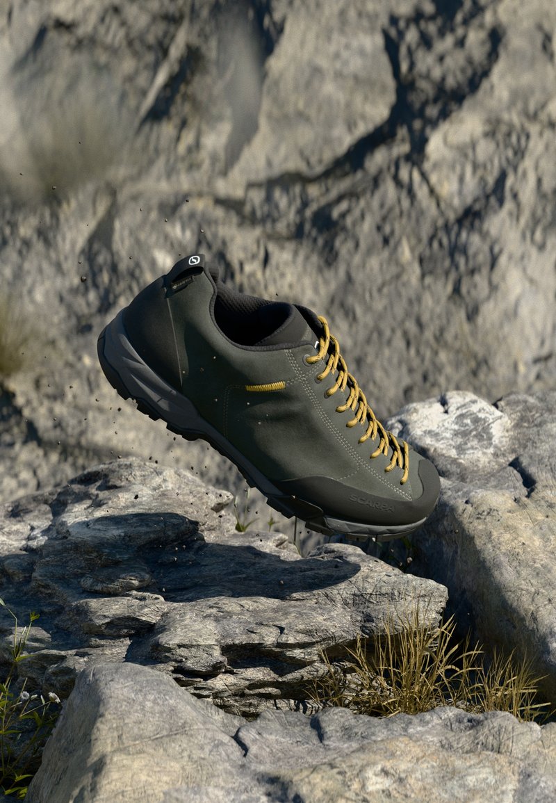 Green outdoor shoe with a textured finish, yellow laces, rubber sole, and a sleek design, positioned above rocky terrain.