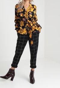 Svart blommig blus med gula och lila blommor, knutet vid midjan, ihop med byxor i svart och vitt rutmönster och glänsande klackboots.