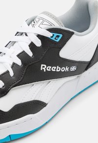 Reebok-sneakers med svart läder och vit syntetisk ovandel, turkosa detaljer och texturerad sula med vita snörningar och logotyp.