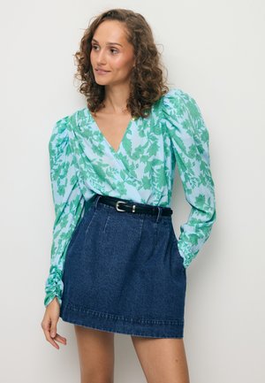 Vrouw met krullend haar, gekleed in een blauwe blouse met groene bladpatronen en opgeblazen mouwen, gecombineerd met een donkere denim rok met ceintuur.