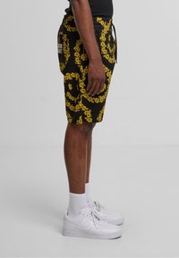 Svarte shorts med gult blomsterdesign, elastisk midje, sidelommer og snor. Kombinert med hvite joggesko og ankelsokker.