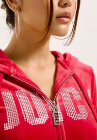 Felpa con zip in velluto rosso, caratterizzata da lettere in strass argento che formano la scritta "JUICY" e da un cursore della zip metallico con il nome del brand. Orecchini a cerchio grandi in evidenza.