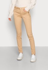 Beige slim-fit byxor med dragkedjefickor, gjorda av mjukt bomullsblandat tyg. Bärs med en ljuströja och vita sneakers.