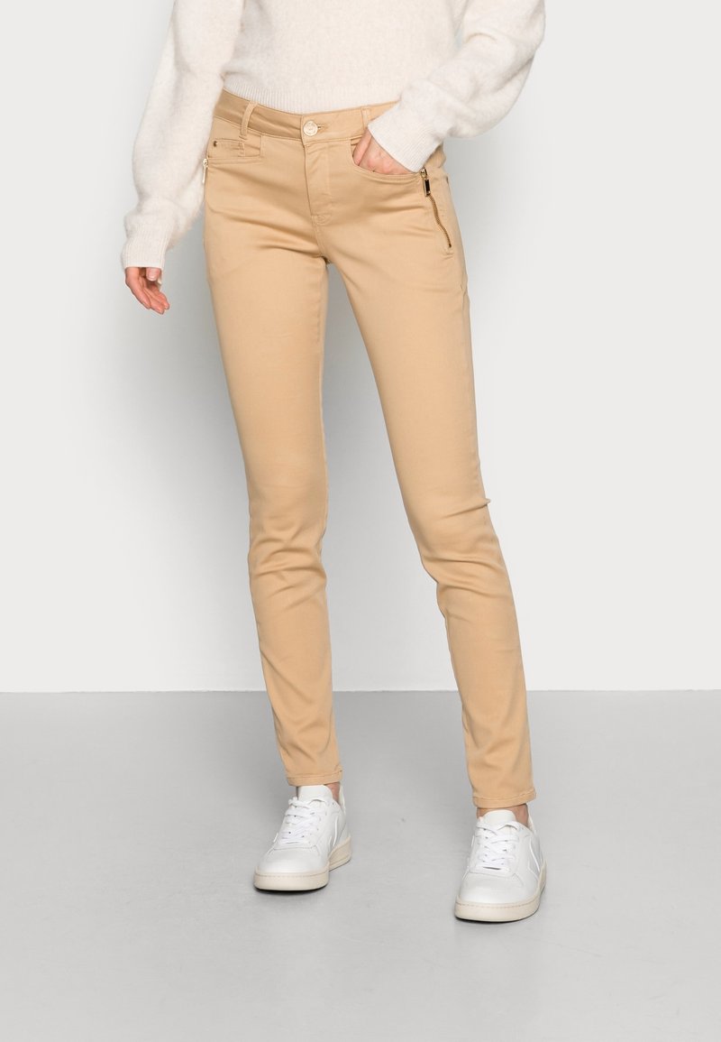 Beige slim-fit byxor med dragkedjefickor, gjorda av mjukt bomullsblandat tyg. Bärs med en ljuströja och vita sneakers.