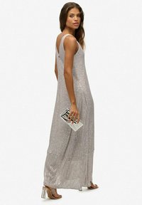 Langes, ärmelloses silbernes Kleid mit V-förmigem Rückendesign, das eine schimmernde Textur aufweist. Das Model hält eine rechteckige metallische Clutch und trägt klare High Heels.