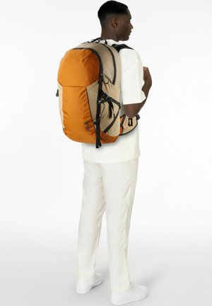 Un homme vêtu d'une tenue blanche est debout de côté avec un grand sac à dos de randonnée orange et beige fixé par des sangles de poitrine et de taille.