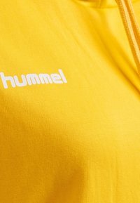 Sudadera amarilla con una textura suave y un logo "hummel" blanco. Presenta una capucha con cordón y un diseño informal de tipo pullover.