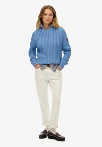 Pull en maille torsadée bleu clair avec un col rond, superposé sur une chemise rayée ; associé avec un pantalon crème et des mocassins marron.