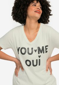 T-shirt a maniche corte e scollatura a V in crema chiaro con testo in paillettes nere "YOU♥ME OUI." Tessuto morbido con una vestibilità comoda.