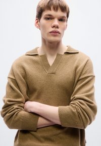 Pull en maille beige avec col en V, large, et épaules tombantes. Le matériau semble doux avec une texture subtile. Le mannequin pose les bras croisés.