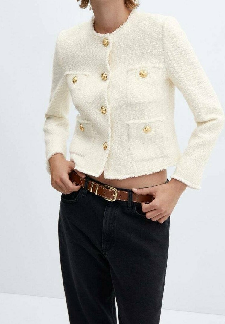 Femme portant une veste crème texturée avec des boutons dorés et quatre poches avant, associée à un pantalon noir et une ceinture marron.