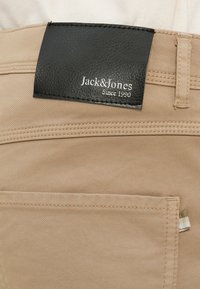 Μπεζ παντελόνι από βαμβάκι με μαύρη δερμάτινη ετικέτα στη μέση που γράφει «Jack & Jones Since 1990». Περιλαμβάνει κανονική ραφή και πίσω τσέπη.